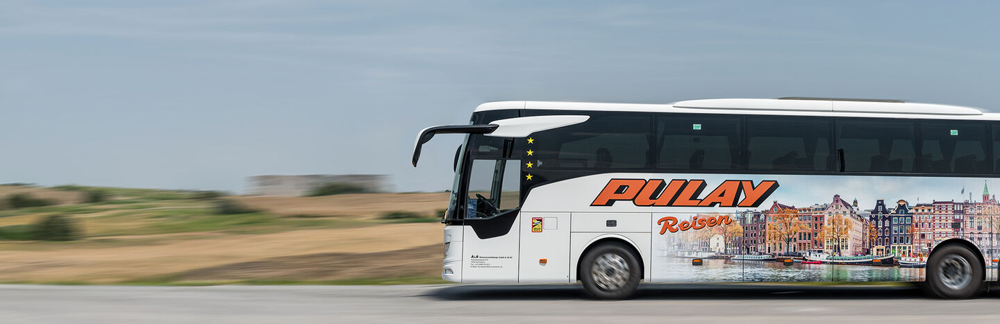 Ein weißer Pulay-Reisen-Reisebus mit Stadtmotiven fährt auf einer Landstraße mit Feldern im Hintergrund.