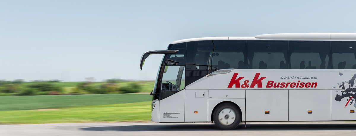 Ein weißer Reisebus von K&K Busreisen fährt auf einer Straße mit grünen Feldern im Hintergrund.