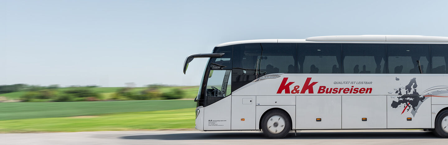 Ein weißer Reisebus von K&K Busreisen fährt auf einer Straße mit grünen Feldern im Hintergrund.
