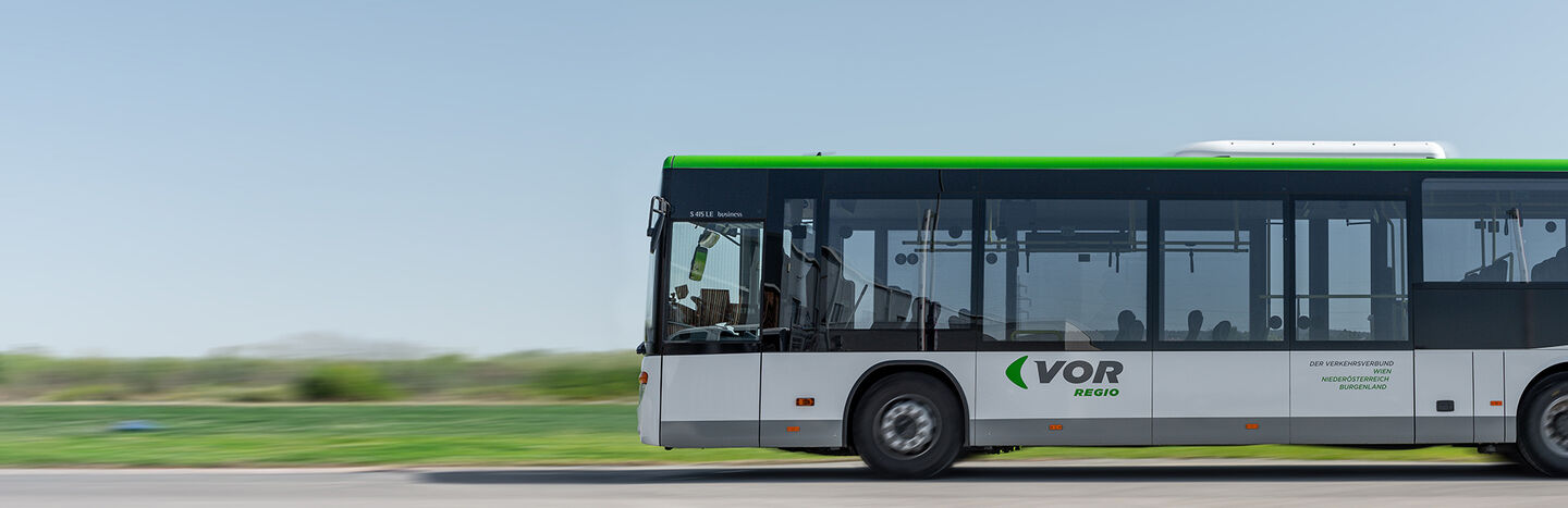 Ein weiß-grüner VOR-Regio-Stadtbus fährt auf einer Straße mit Gras und Bäumen im Hintergrund.