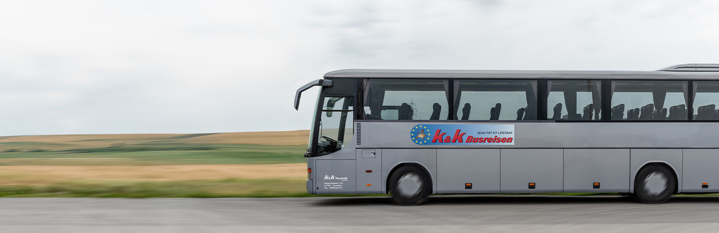 Ein silberner Reisebus fährt auf einer Landstraße mit Feldern und einem bewölkten Himmel im Hintergrund.