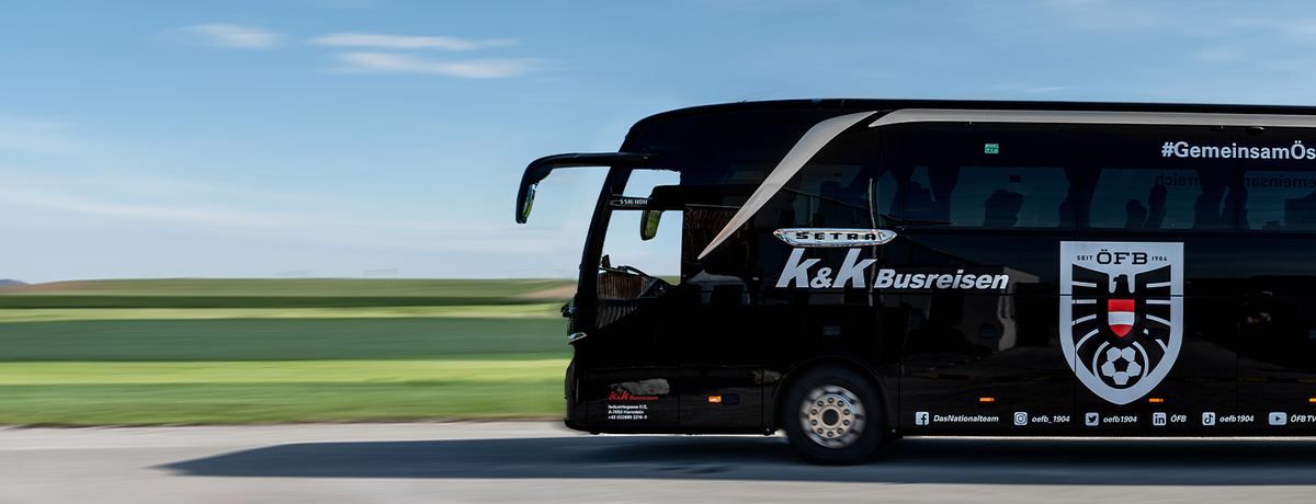 Ein schwarzer Mannschaftsbus mit Logos fährt auf einer Straße neben grünen Feldern unter blauem Himmel.
