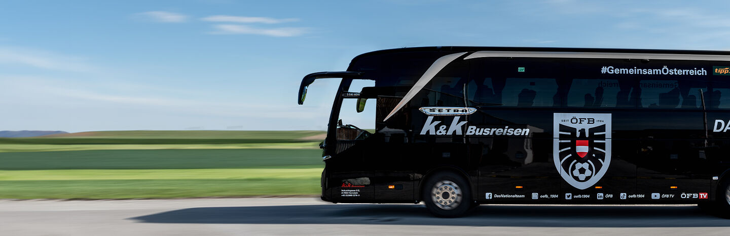 Ein schwarzer Mannschaftsbus mit Logos fährt auf einer Straße neben grünen Feldern unter blauem Himmel.