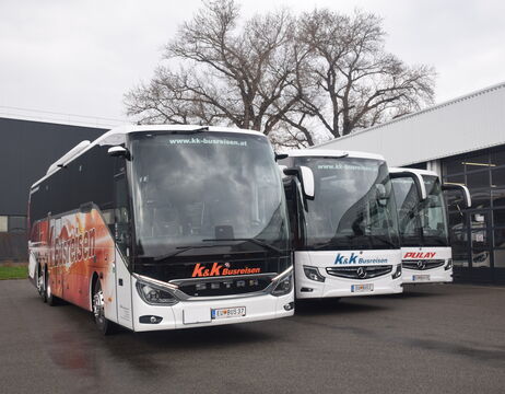 Drei moderne Reisebusse, die nebeneinander vor einem Gebäude geparkt sind, mit Bäumen im Hintergrund.