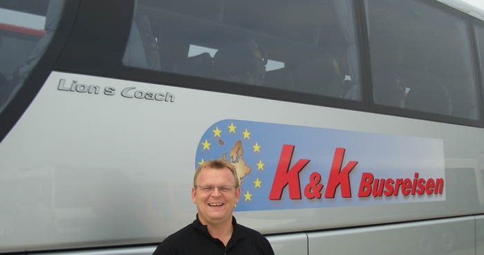 Ein lächelnder Mann steht vor einem Reisebus von K&K Busreisen mit einem Euro-Logo.