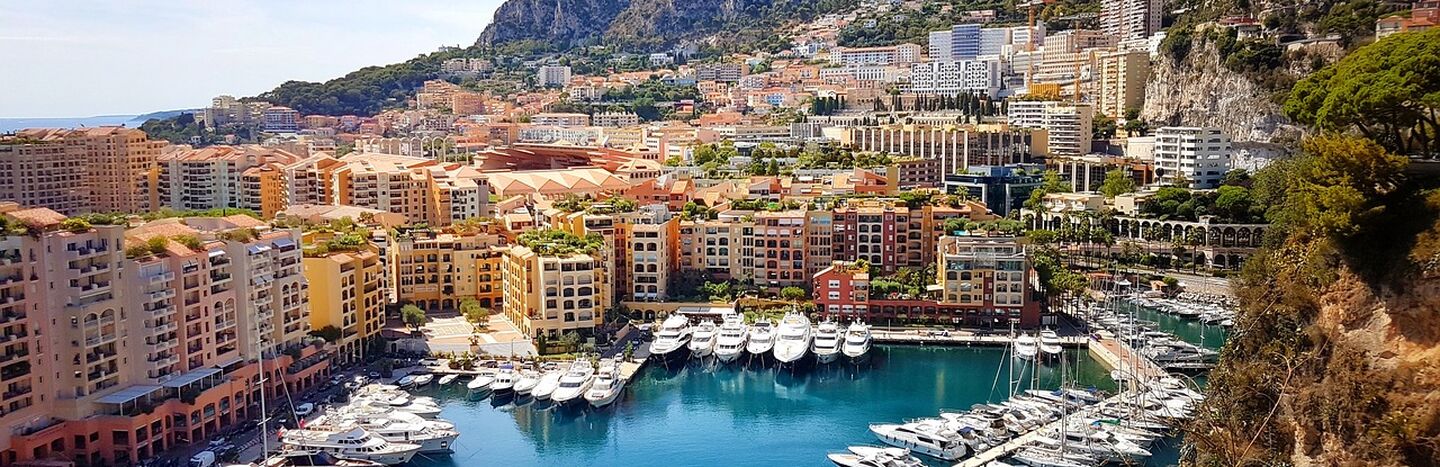 Ein Jachthafen mit Yachten, bunten Gebäuden und Bergen im Hintergrund unter blauem Himmel.