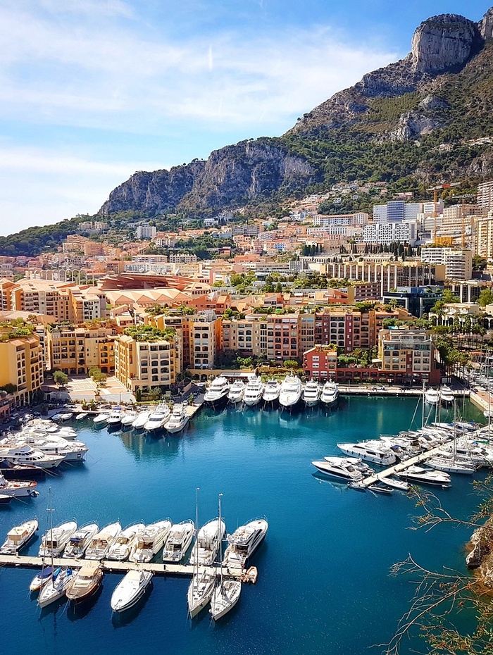 Ein Jachthafen mit Yachten, bunten Gebäuden und Bergen im Hintergrund unter blauem Himmel.