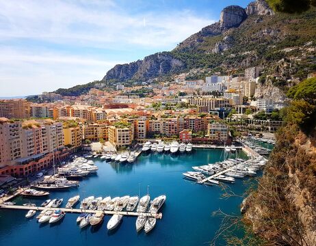 Ein Jachthafen mit Yachten, bunten Gebäuden und Bergen im Hintergrund unter blauem Himmel.