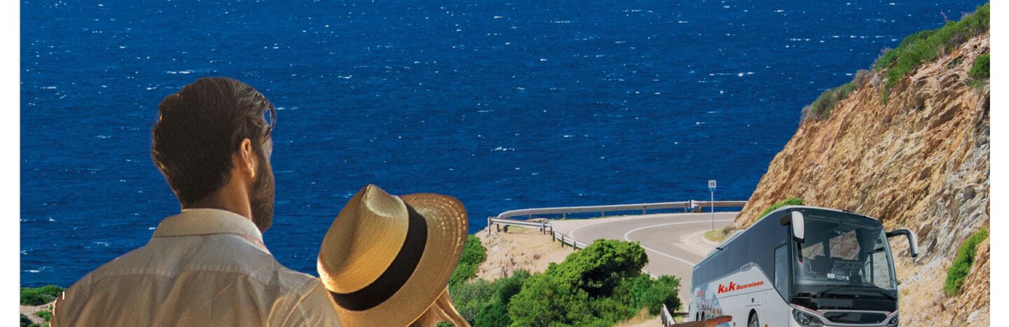 Ein Paar schaut auf einen Bus, der eine Küstenstraße entlangfährt, mit blauem Meer und Bergen im Hintergrund.