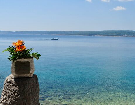 Ein Segelboot schwimmt auf ruhigem, blauem Wasser mit entfernten Hügeln und einer Steinmauer im Vordergrund.