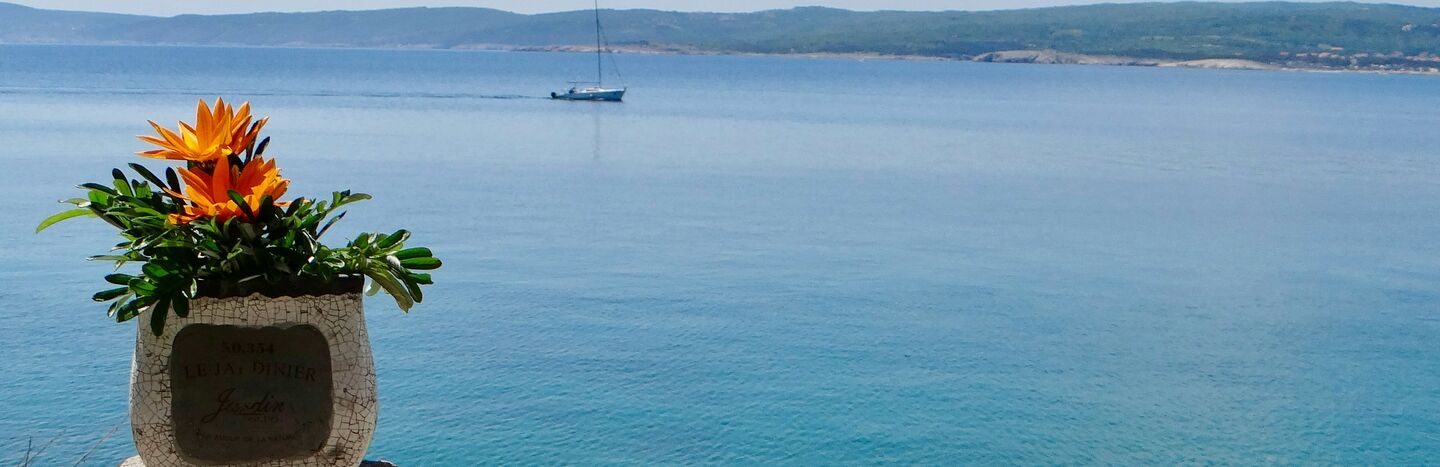 Ein Segelboot schwimmt auf ruhigem, blauem Wasser mit entfernten Hügeln und einer Steinmauer im Vordergrund.