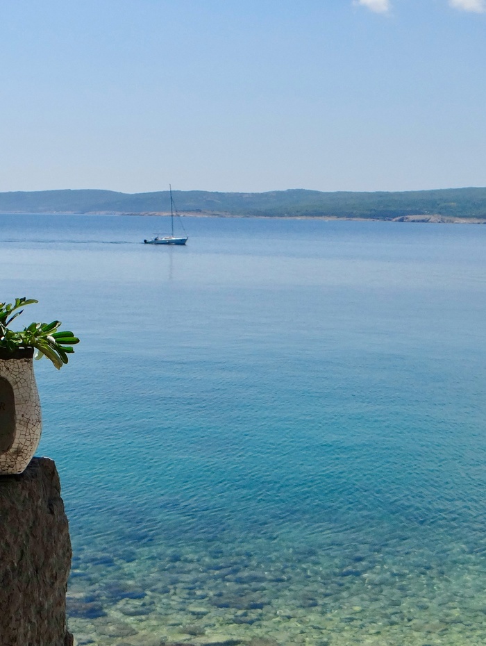 Ein Segelboot schwimmt auf ruhigem, blauem Wasser mit entfernten Hügeln und einer Steinmauer im Vordergrund.