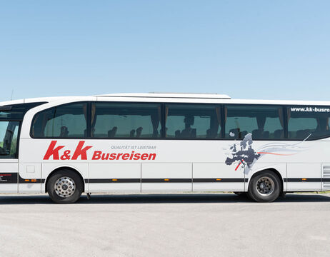 Ein weißer Reisebus von K&K Busreisen parkt unter blauem Himmel auf dem Bürgersteig.