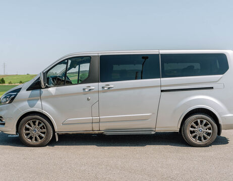 Ein silberner Lieferwagen, der auf einer asphaltierten Straße geparkt ist, mit einem grasbewachsenen Feld und einem klaren Himmel im Hintergrund.