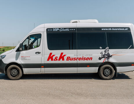 Weißer K&K Busreisen VIP-Shuttle-Kleinbus mit Logo und Website an der Seite, der auf einer Straße geparkt ist.