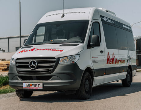 Weißer Mercedes-Benz-Kleinbus mit k&k Busreisen-Branding, geparkt an einer sonnigen Straße.