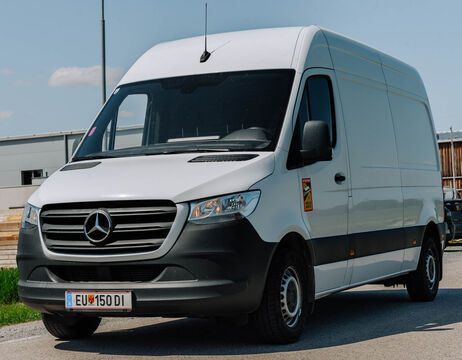 Ein weißer Mercedes-Benz Sprinter parkt auf einer Straße mit Gebäuden im Hintergrund.
