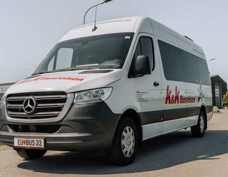 Ein weißer Mercedes-Benz Kleinbus mit dem Schriftzug k&k Busreisen parkt an einer sonnigen Straße.