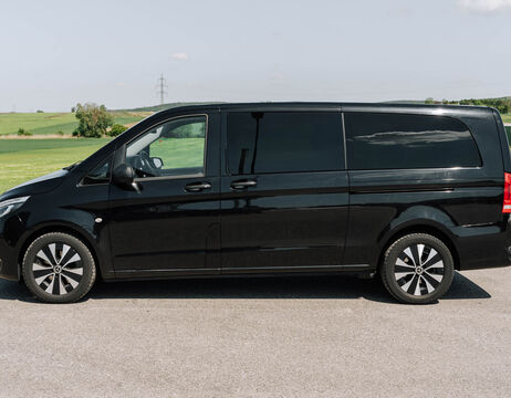 Ein schwarzer Lieferwagen parkt auf einer gepflasterten Straße mit grünen Feldern und Hügeln im Hintergrund.