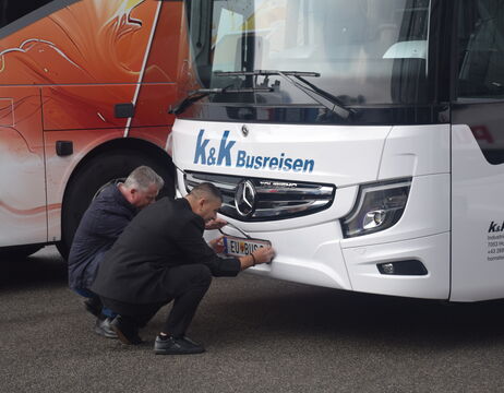 Zwei Männer knien vor einem weißen Bus und befestigen bzw. überprüfen das vordere Nummernschild.