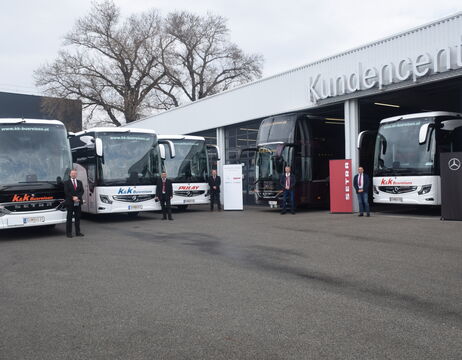 Fünf Personen stehen an einem bewölkten Tag neben vier großen Bussen vor einem Gebäude mit der Aufschrift Kundencenter.