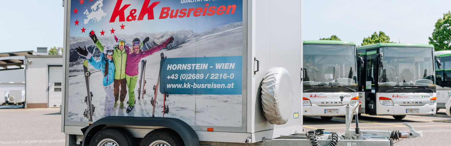 Ein Anhänger mit einer Skireise-Werbung neben grünen und weißen Bussen auf einem Parkplatz.