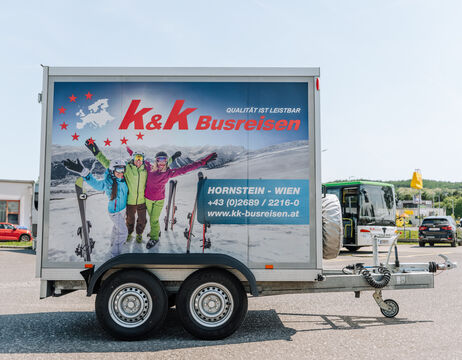 Werbeanhänger mit drei lächelnden Skifahrern und dem Logo von K&K Busreison, geparkt auf einem Parkplatz.