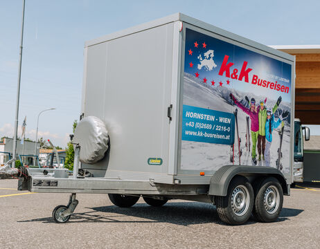 Ein grauer Anhänger mit k&k Busreisen und einer Gruppe von Skifahrern, die auf der Seite abgebildet sind, ist im Freien geparkt.
