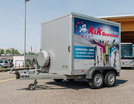 Grauer Anhänger mit k&k Busreisen-Werbung, auf dem Menschen in Skiausrüstung abgebildet sind, geparkt auf einem Parkplatz.