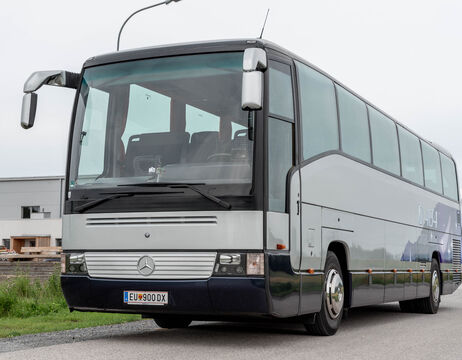 Ein silberner Mercedes-Benz Reisebus, der am Straßenrand geparkt ist, mit Gebäuden im Hintergrund.