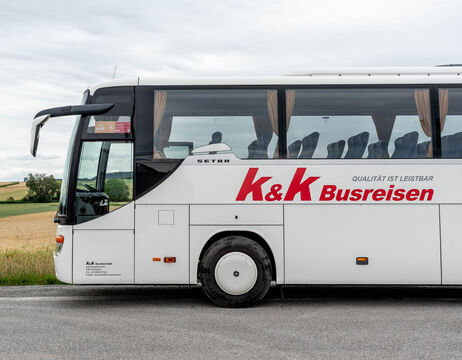 Weißer Reisebus mit K&K Busreisen-Logo, geparkt auf einer Landstraße, Felder und Himmel im Hintergrund.