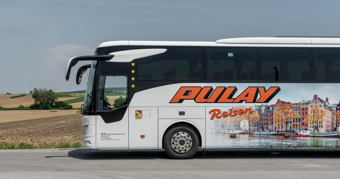 Ein weißer Pulay Reisen Reisebus parkt auf einer Straße mit Feldern und einem bewölkten Himmel im Hintergrund.