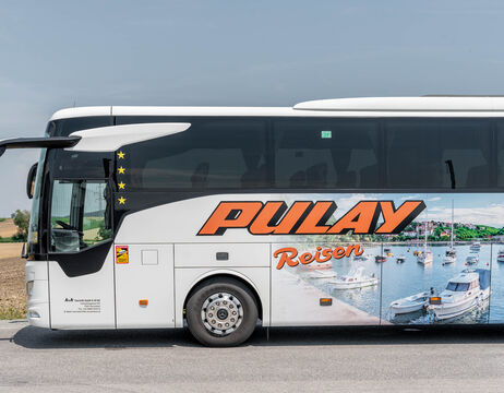 Ein weißer Reisebus mit der Aufschrift Pulay Reisen und Bildern von Booten an der Seite, der an einer Straße geparkt ist.