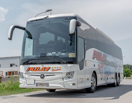 Ein weißer Pulay-Reisebus mit Mercedes-Benz-Logo parkt auf einer asphaltierten Straße unter freiem Himmel.
