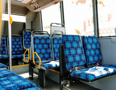 Reihen von leeren, blau gemusterten Sitzen in einem gut beleuchteten Stadtbus, durch dessen Fenster das Sonnenlicht scheint.