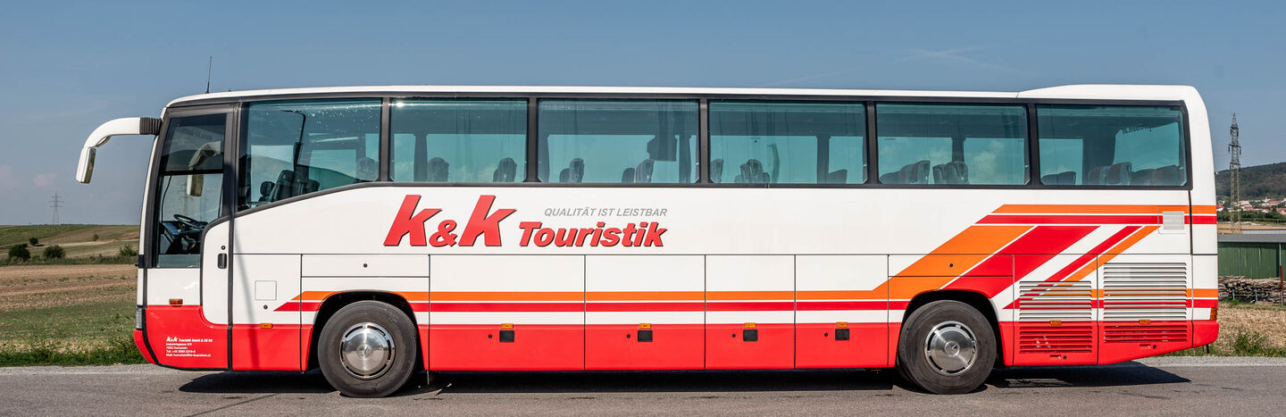 Ein weiß-roter Reisebus mit der Aufschrift K&K Touristik parkt auf einer Landstraße mit Feldern im Hintergrund.