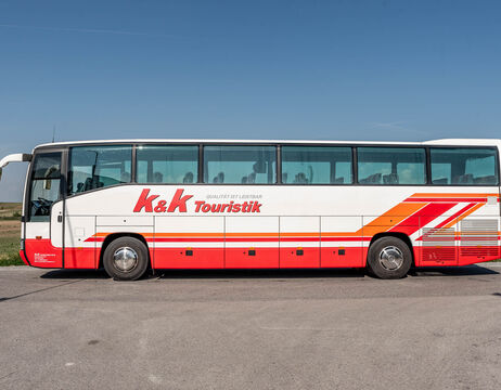 Ein weiß-roter Reisebus mit der Aufschrift K&K Touristik parkt auf einer Landstraße mit Feldern im Hintergrund.