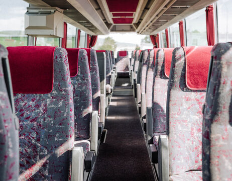Leere Bussitze mit roten und grauen Polstern, ein Gang in der Mitte, helles Tageslicht durch die Fenster.