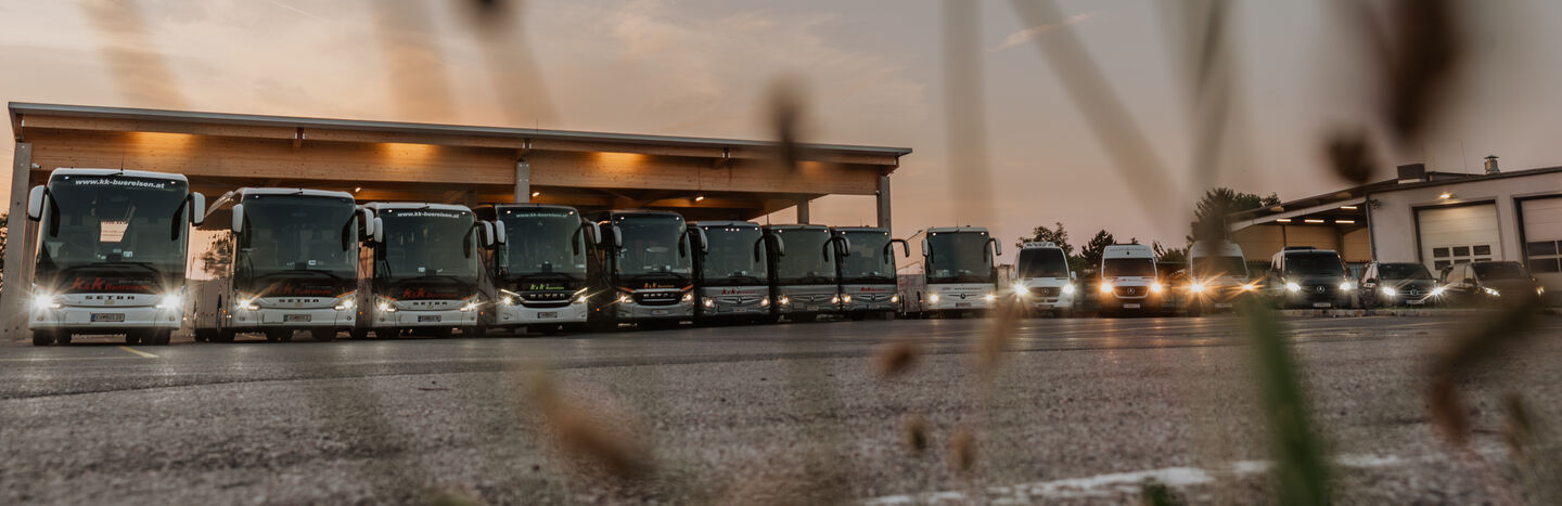 Eine Reihe von Bussen und Lieferwagen, die in der Dämmerung nebeneinander vor einem Gebäude geparkt sind, mit eingeschalteten Scheinwerfern.