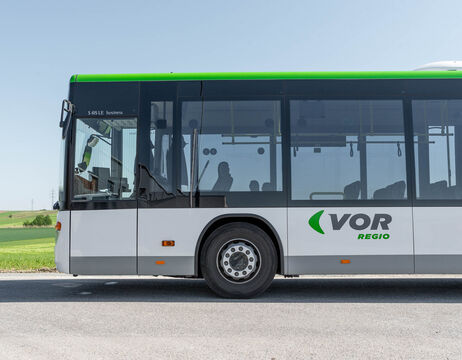 Seitenansicht eines weiß-grünen VOR-Regio-Busses, der an einer Straße mit einer Wiese im Hintergrund geparkt ist.