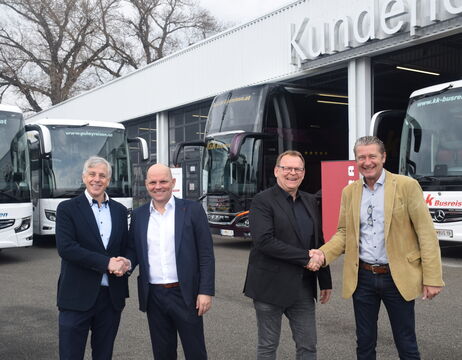 Vier Männer in Geschäftskleidung schütteln sich die Hände und lächeln vor mehreren Bussen vor einem Gebäude.