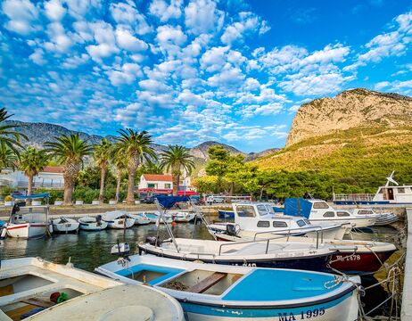 Boote in einem Jachthafen, Palmen, Berge und ein strahlend blauer Himmel mit vereinzelten Wolken.