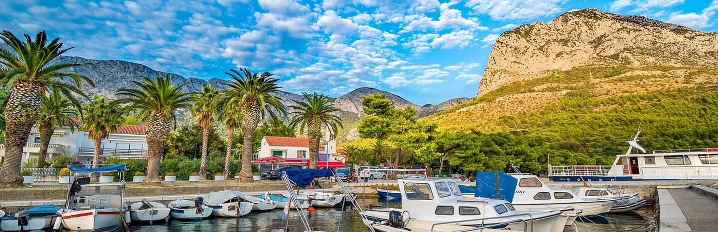 Boote in einem Jachthafen, Palmen, Berge und ein strahlend blauer Himmel mit vereinzelten Wolken.