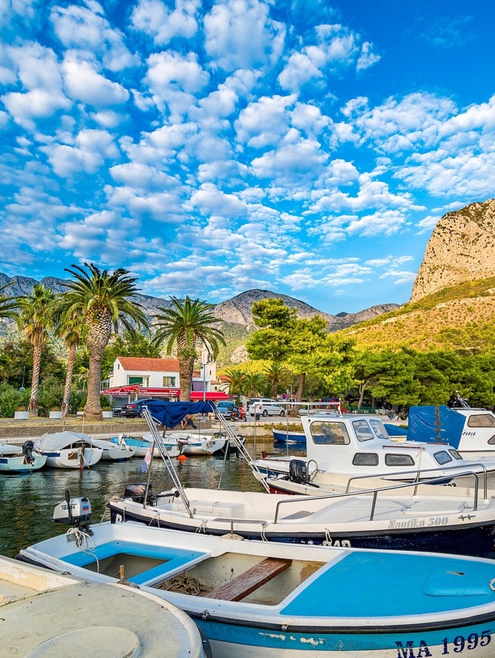 Boote in einem Jachthafen, Palmen, Berge und ein strahlend blauer Himmel mit vereinzelten Wolken.