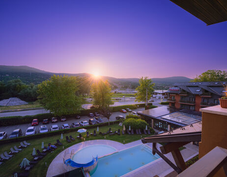Hotelpool und Liegestühle bei Sonnenuntergang, mit Bergen und parkenden Autos im Hintergrund.
