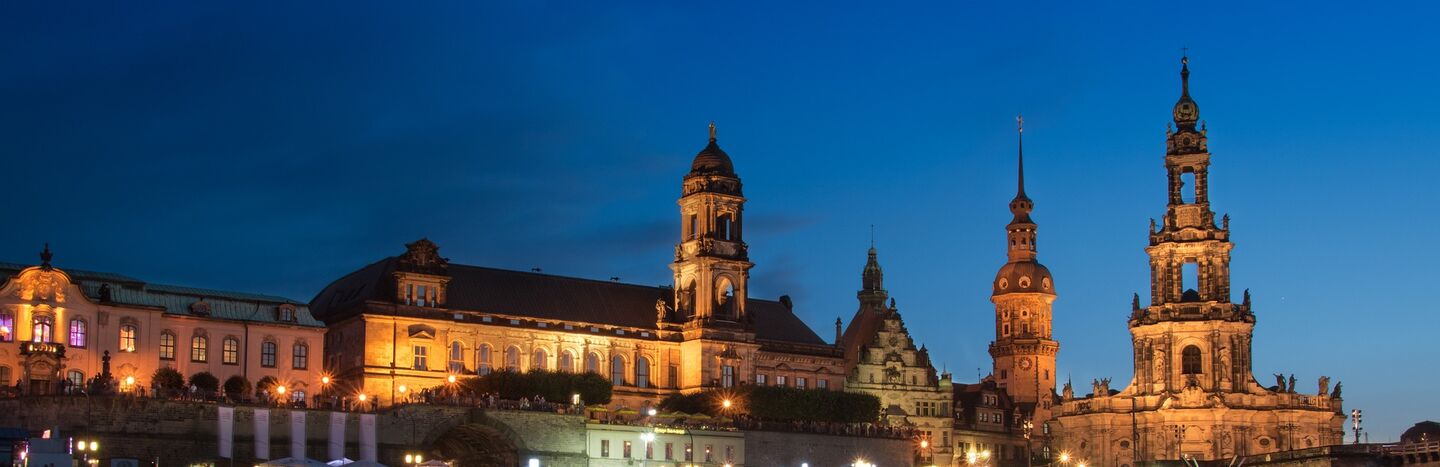Bunte historische Gebäude und eine Kirche mit grüner Kuppel spiegeln sich in einem ruhigen Fluss unter einem bewölkten Himmel.