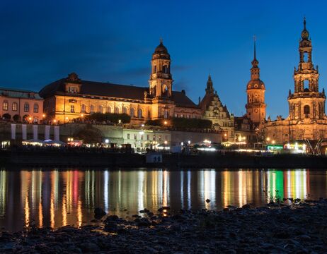 Bunte historische Gebäude und eine Kirche mit grüner Kuppel spiegeln sich in einem ruhigen Fluss unter einem bewölkten Himmel.