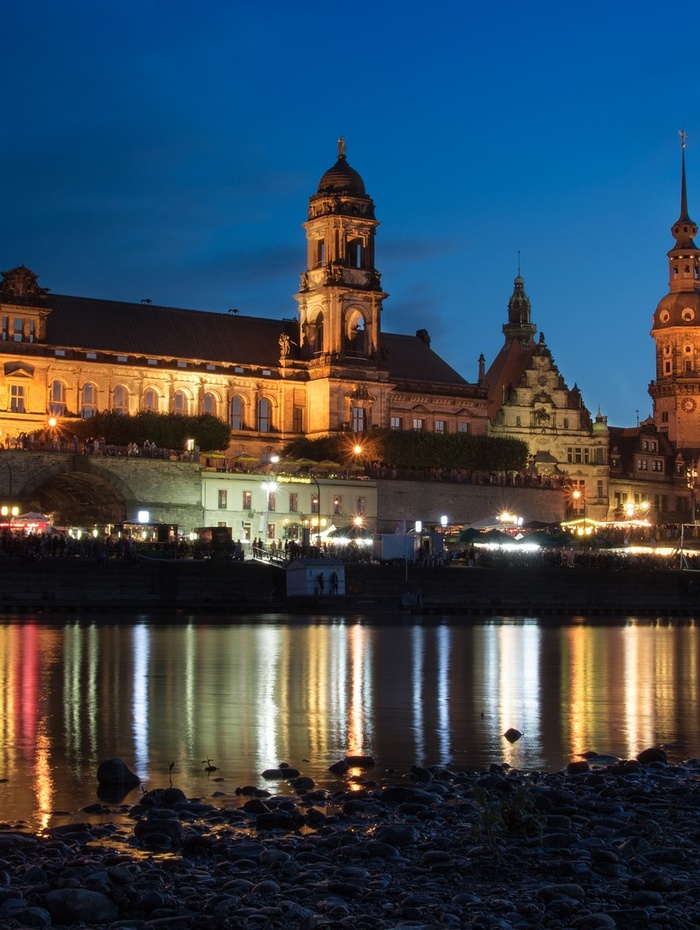 Bunte historische Gebäude und eine Kirche mit grüner Kuppel spiegeln sich in einem ruhigen Fluss unter einem bewölkten Himmel.