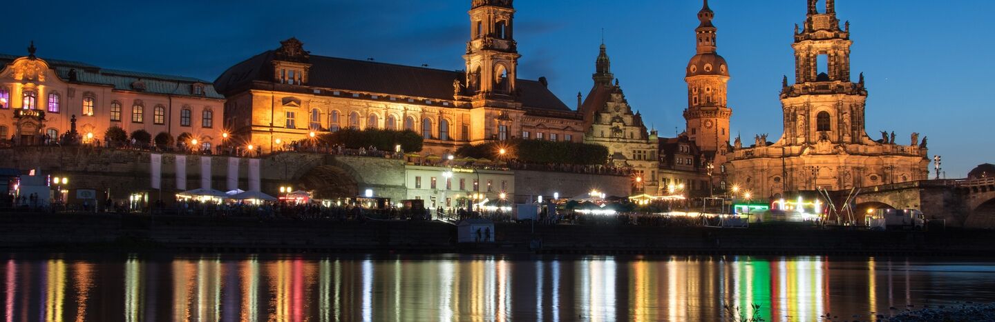 Bunte historische Gebäude und eine Kirche mit grüner Kuppel spiegeln sich in einem ruhigen Fluss unter einem bewölkten Himmel.