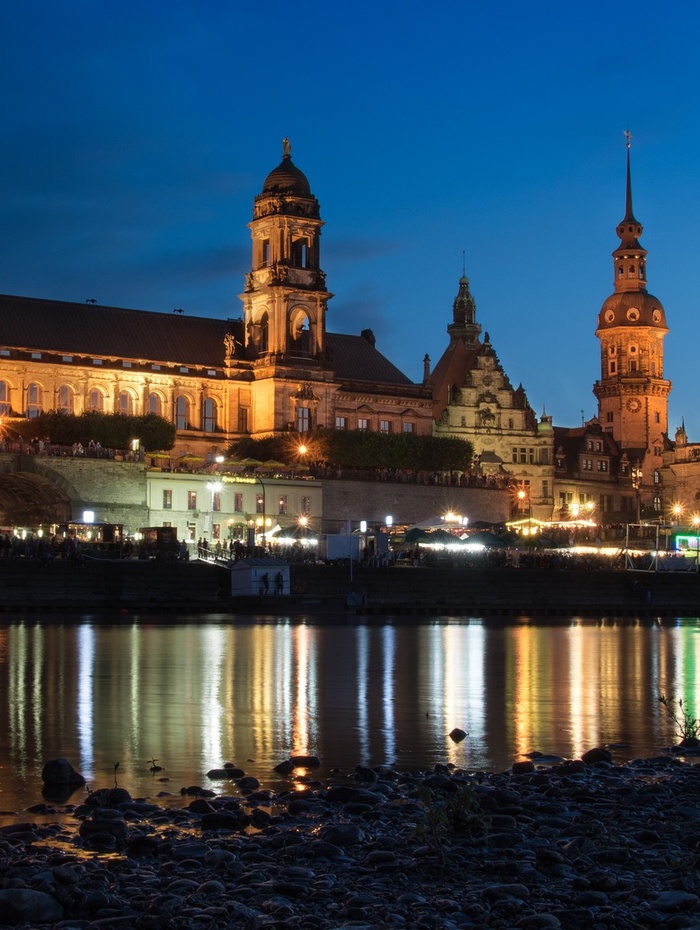 Bunte historische Gebäude und eine Kirche mit grüner Kuppel spiegeln sich in einem ruhigen Fluss unter einem bewölkten Himmel.